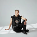 Man sitting in minimalist room wearing black sleeveless shirt with compass graphic, black pants and black shoes